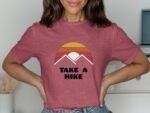 Woman wearing a pink t-shirt with a sunset and mountain graphic, text reads "TAKE A HIKE." Background is simple and light.