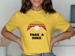 A woman wearing a yellow shirt with a mountain graphic and the text "TAKE A HIKE," posing in front of a light background.