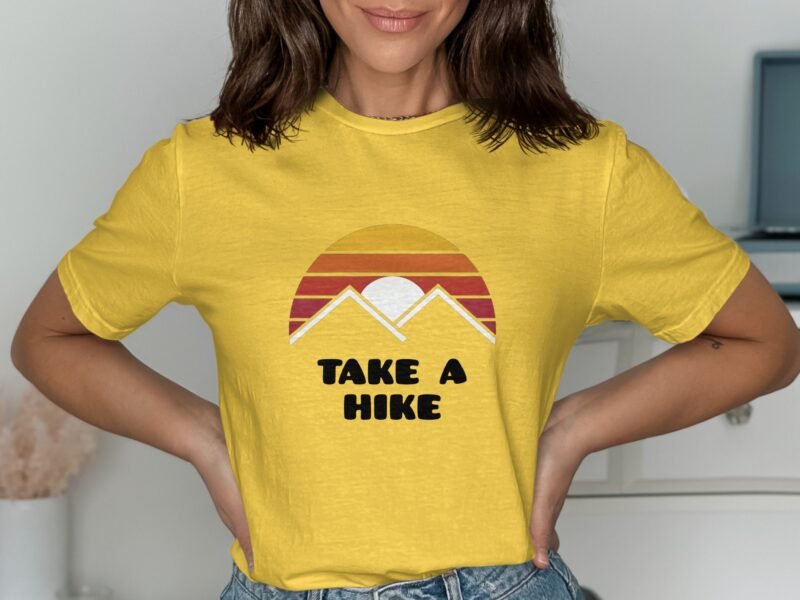 A woman wearing a yellow shirt with a mountain graphic and the text "TAKE A HIKE," posing in front of a light background.