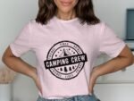 Woman in a pink t-shirt with a “Camping Crew” logo, featuring a tent and trees, standing in front of a simple background.