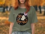 A person wearing an olive green t-shirt featuring a ghost graphic with "30 & Bootiful" text and Halloween-themed decorations.