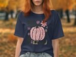 A woman with red hair wears a navy shirt featuring a pink pumpkin graphic among colorful flowers, set in an autumn landscape.
