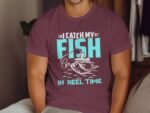 A person wearing a maroon t-shirt with the text "I CATCH MY FISH IN REEL TIME" and a fishing graphic.