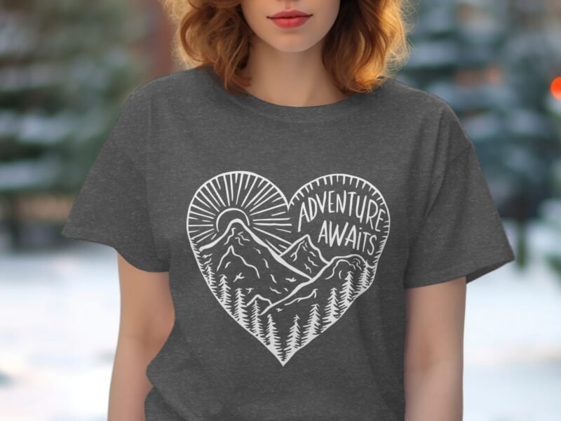 A woman wearing a gray t-shirt with a heart design featuring mountains, trees, and the text "Adventure Awaits."