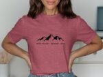 A woman poses in a pink t-shirt with "Hike More - Worry Less" and mountain graphics printed on the front.