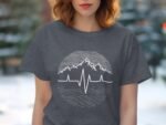 A woman with wavy hair wears a gray t-shirt featuring a mountain and heartbeat graphic against a blurred outdoor background.