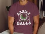 A man in a maroon t-shirt featuring the text "Easily Distracted By Balls" and a golf graphic.