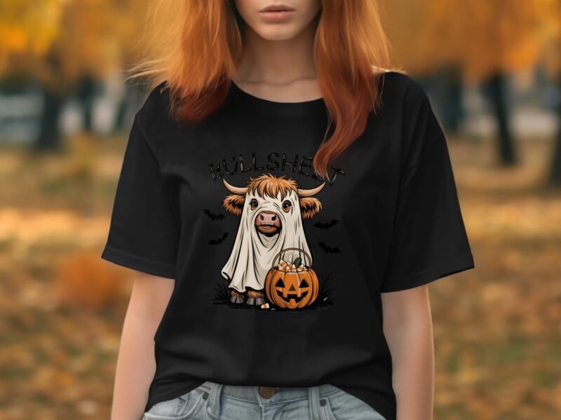 A woman in a black t-shirt featuring a cartoon cow in a ghost costume holding a pumpkin, with autumn leaves in the background.