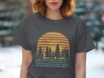 A woman wearing a gray t-shirt with a sunset and pine tree graphic stands outdoors.