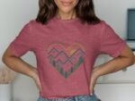 A woman wearing a pink t-shirt with a heart design featuring mountains and trees, smiling against a neutral background.