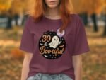 A woman stands outdoors in a burgundy t-shirt featuring a ghost and the text "30 & Bootiful" with Halloween motifs.