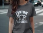 Woman wearing a gray t-shirt featuring the text "PISTON BROKE" with graphic elements, standing on a city street.