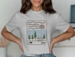 A woman wearing a gray t-shirt featuring an illustrated camping scene with trees, a caravan, water, and a sun.