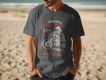 A man wearing a dark gray t-shirt with motorcycle graphics stands on a beach, sunlit with ocean in the background.