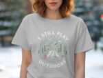A woman in a gray t-shirt displays a graphic featuring mountains, trees, and the text "I Still Play Outdoors."