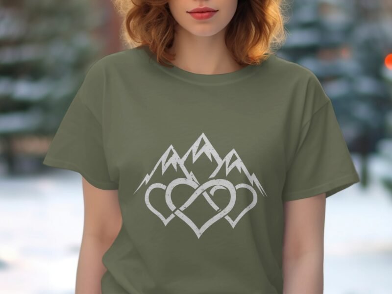 A woman wearing an olive green t-shirt with mountain and heart graphic, in a snowy outdoor setting.