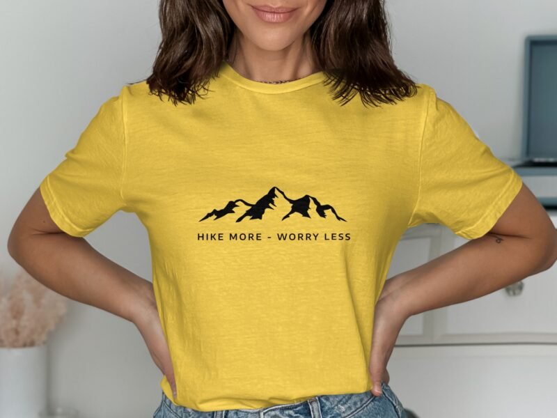A woman in a yellow t-shirt with "Hike More - Worry Less" text and mountain graphics, standing indoors.