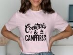 A woman wears a light pink t-shirt with "Cocktails & Campfires" printed in bold black letters.