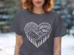 A woman with wavy hair wears a dark gray t-shirt featuring a heart graphic with mountains and text reading "ADVENTURE AWAITS."