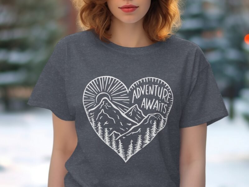 A woman with wavy hair wears a dark gray t-shirt featuring a heart graphic with mountains and text reading "ADVENTURE AWAITS."