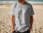 Man wearing a gray t-shirt with barber-themed graphics standing on a beach, with water and sand in the background.