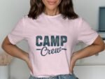 A woman wearing a pink t-shirt with the text "CAMP Crew" in bold blue letters, smiling and posing stylishly.