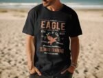 Man wearing a black t-shirt with "EAGLE" graphic and text on a sandy beach with ocean in the background.