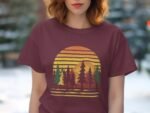 Woman wearing a burgundy t-shirt with a sunset and forest graphic design, outdoors with blurred trees in the background.