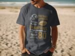 A man stands on a sandy beach wearing a gray t-shirt featuring football-themed graphics and text.