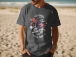 A man in a dark gray T-shirt with a colorful surf and palm tree graphic stands on a sandy beach by the ocean.