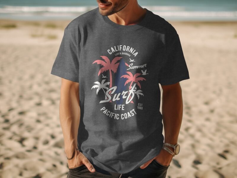 A man in a dark gray T-shirt with a colorful surf and palm tree graphic stands on a sandy beach by the ocean.
