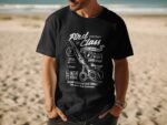 Man wearing a black t-shirt with barbershop-themed graphic and text, standing on a sandy beach with ocean in background.
