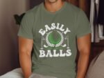A man wearing an olive green t-shirt with the phrase “EASILY Distracted By BALLS” and a golf-themed graphic.