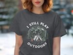 A woman with wavy hair wears a dark t-shirt featuring the text "I STILL PLAY OUTDOORS" and a camping scene design.