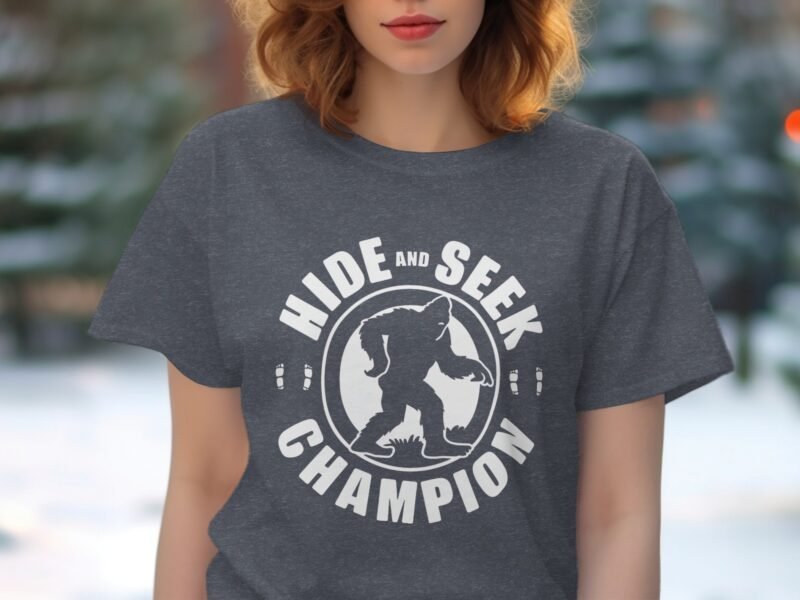 A woman with wavy hair wearing a gray t-shirt featuring a "Hide and Seek Champion" design with a Bigfoot illustration.
