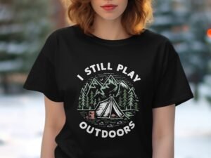 A woman wearing a black T-shirt featuring the text "I STILL PLAY OUTDOORS" with a camping design and trees.