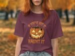 A woman in a maroon t-shirt featuring a pumpkin graphic and the text "If you've got it, haunt it," against a fall background.