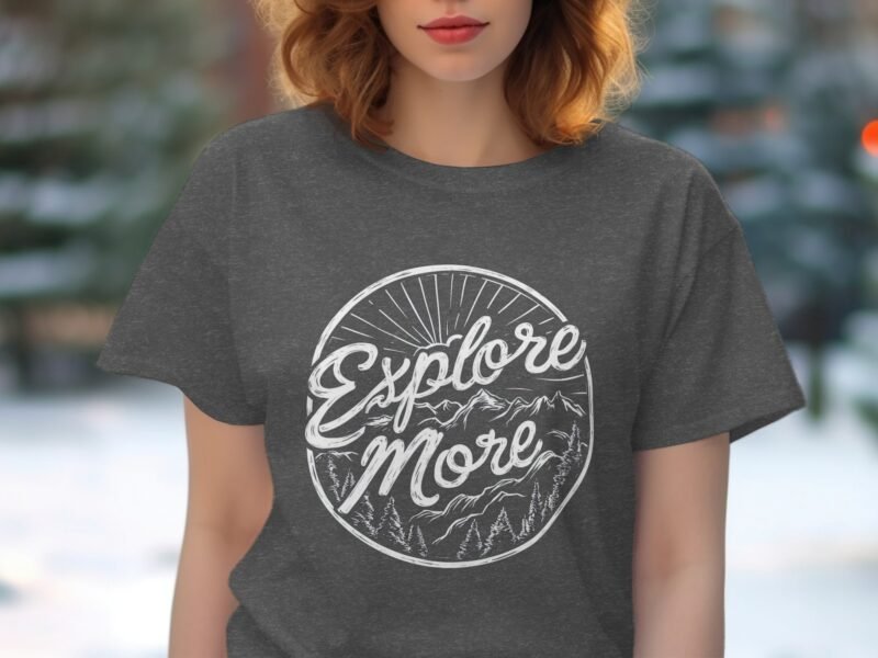 Woman wearing a gray t-shirt with the phrase "Explore More" and mountain graphic, outdoors with blurred greenery in background.