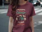 A person wearing a maroon t-shirt featuring a camping-themed graphic with mountains, a tent, and adventure text.