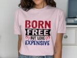 A woman wearing a pink t-shirt with the text "BORN FREE BUT LOVE EXPENSIVE" in bold, colorful letters.
