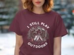 A woman wearing a maroon t-shirt with "I STILL PLAY OUTDOORS" and a camping graphic featuring mountains and a tent.