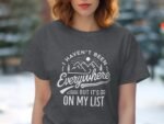 Woman wearing a dark gray t-shirt with a travel-themed quote and mountain graphic, set against a blurred outdoor background.