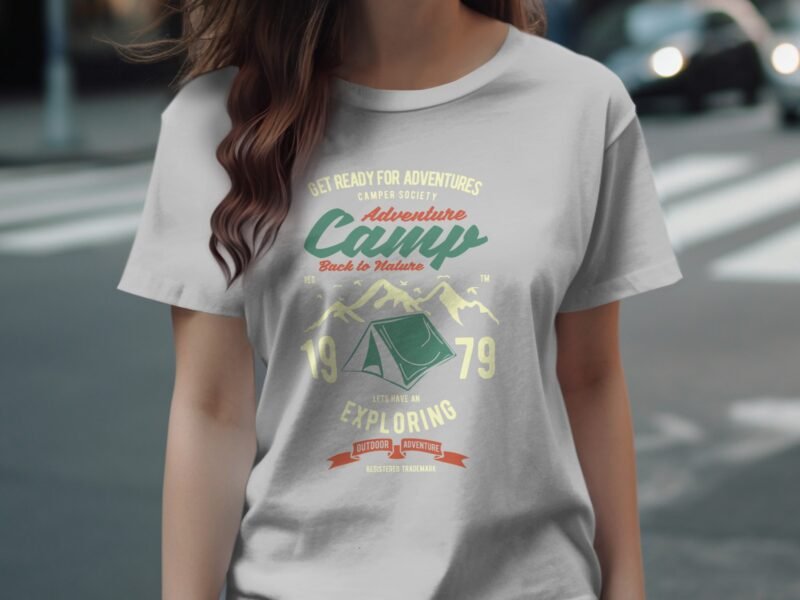 A woman wearing a light gray t-shirt with a vintage camping graphic featuring mountains, a tent, and adventure-themed text.