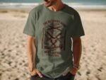 A man wearing a green t-shirt with a retro butchery graphic stands on a beach with waves in the background.