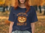 Person wearing a navy t-shirt with a pumpkin graphic and text: "If you've got it, haunt it," in an autumn setting.