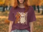 A person with red hair wears a maroon t-shirt featuring a ghost design with flowers against a blurred autumn background.