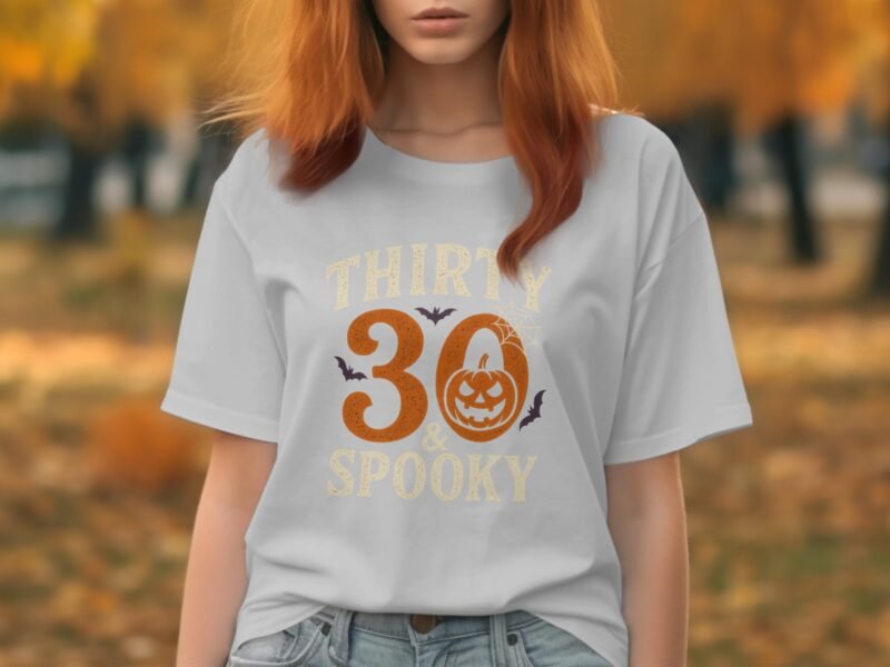 Woman with shoulder-length red hair wears a white t-shirt featuring "Thirty & Spooky" graphic, set against autumn foliage.