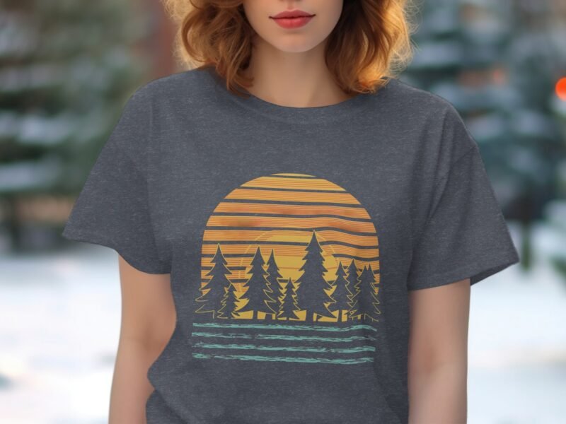 A woman wearing a gray t-shirt with a sunset and trees graphic stands in a blurred outdoor setting.