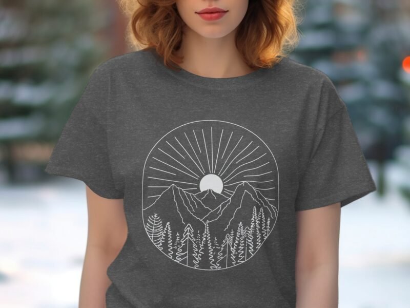 A woman in a gray t-shirt with a white mountain and sun graphic, surrounded by blurred outdoor greenery.
