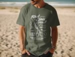 Man wearing a green t-shirt with barbershop-themed graphics and text, standing on a beach.
